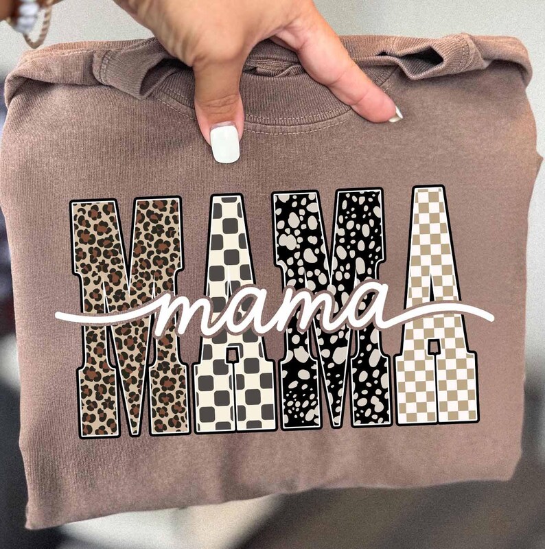May include: A brown sweatshirt with the word "MAMA" in large, decorative letters. The letters have various patterns, including leopard print, checkered, and speckled designs. The word "mama" is written in cursive below.