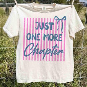 Puede incluir: Camiseta color crema con el texto azul "JUST ONE MORE Chapter" sobre un fondo de rayas rosas. Un lazo azul est&aacute; a la derecha. La camiseta est&aacute; colgada en un tendedero.