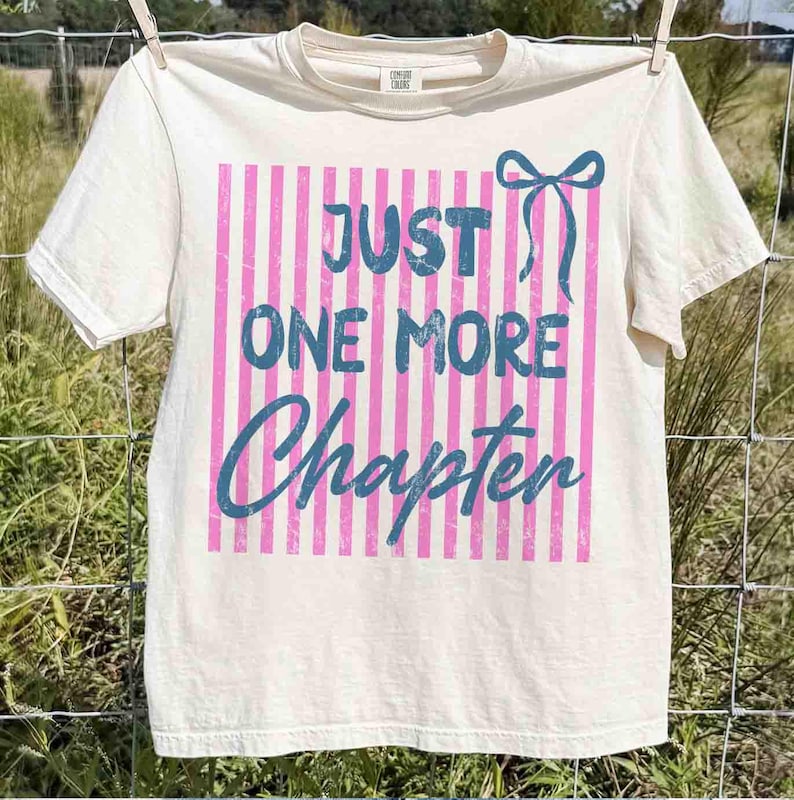 Puede incluir: Camiseta color crema con el texto azul "JUST ONE MORE Chapter" sobre un fondo de rayas rosas. Un lazo azul est&aacute; a la derecha. La camiseta est&aacute; colgada en un tendedero.