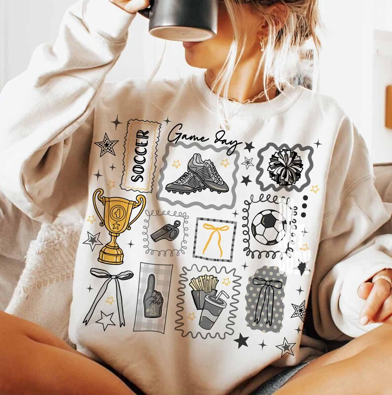 May include: Cream-coloured sweatshirt with a soccer-themed graphic. The design includes a trophy, football, boots, pom-pom, and the words "Game Day" and "Soccer". The graphic is in black, grey, and gold.