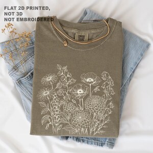 May include: Olive green t-shirt with a white floral design, folded on top of blue jeans. A gold necklace with a small pendant rests on the shirt. The text "FLAT 2D PRINTED, NOT 3D, NOT EMBROIDERED" is visible.