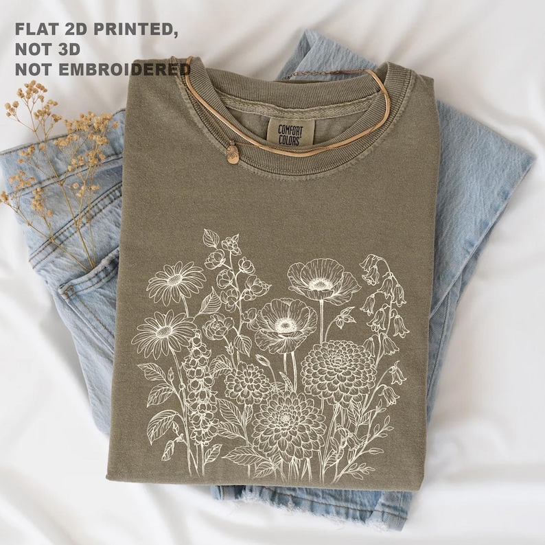 May include: Olive green t-shirt with a white floral design, folded on top of blue jeans. A gold necklace with a small pendant rests on the shirt. The text "FLAT 2D PRINTED, NOT 3D, NOT EMBROIDERED" is visible.