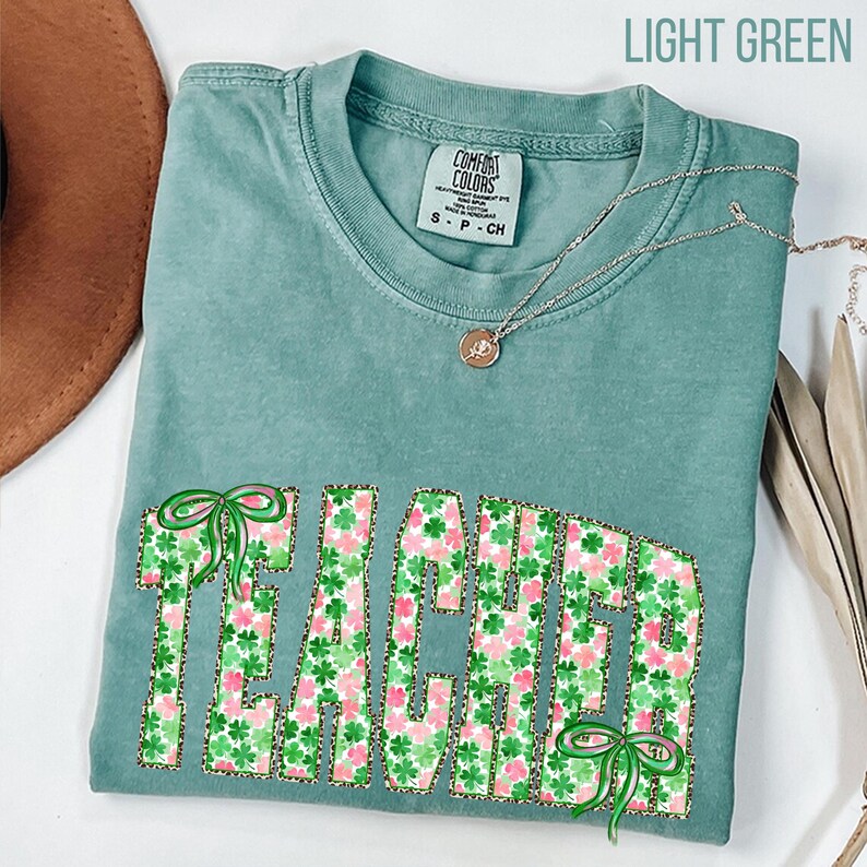 May include: A light green t-shirt with the word "TEACHER" in a floral pattern with green bows. The shirt has a crew neck and short sleeves. A silver necklace with a pendant is draped across the shirt.