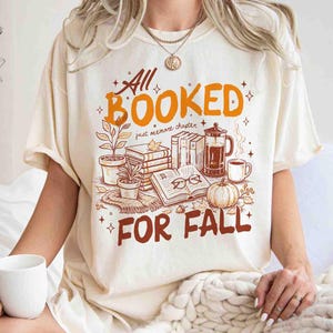 May include: Cream-colored t-shirt with the words "All Booked For Fall" in orange and brown. The graphic includes books, plants, a coffee press, a pumpkin, and eyeglasses. The shirt has short sleeves and a relaxed fit.