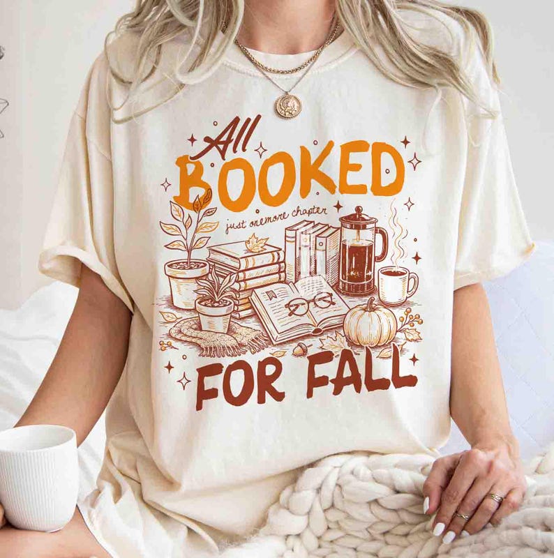 May include: Cream-colored t-shirt with the words "All Booked For Fall" in orange and brown. The graphic includes books, plants, a coffee press, a pumpkin, and eyeglasses. The shirt has short sleeves and a relaxed fit.