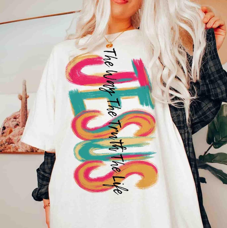 May include: White t-shirt with the word "JESUS" in large, colorful, brushstroke letters. The text "The Way The Truth The Life" is printed vertically on the left side of the word "JESUS". The shirt has short sleeves.