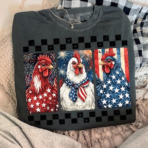 May include: Dark gray sweatshirt featuring three chickens with patriotic designs. One chicken is red with white stars, another is white with a patriotic scarf, and the last is blue with white stars. A black and white checkered pattern is above the chickens.
