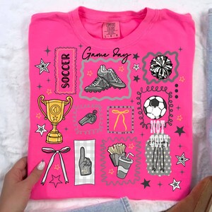 May include: A vibrant pink t-shirt featuring soccer-related illustrations such as football boots, a trophy, and a football. The shirt also has the words "Game Day" and "Soccer", along with star and pom-pom designs.