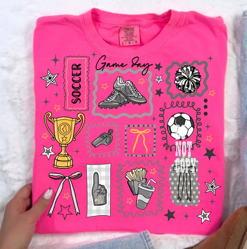 May include: A vibrant pink t-shirt featuring soccer-related illustrations such as football boots, a trophy, and a football. The shirt also has the words "Game Day" and "Soccer", along with star and pom-pom designs.