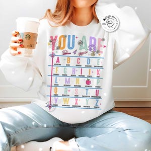 May include: White sweatshirt with a colorful alphabet chart that says "You Are" at the top. Each letter of the alphabet is paired with a positive adjective. For example, "A" is for "Accepting" and "B" is for "Brave".