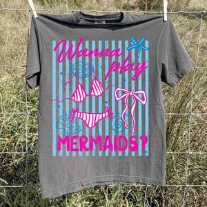 May include: Gray t-shirt with a graphic design featuring a bikini, bow, and starfish, with the text "Wanna Play Mermaids?" in pink and blue. The shirt is hanging on a clothesline.