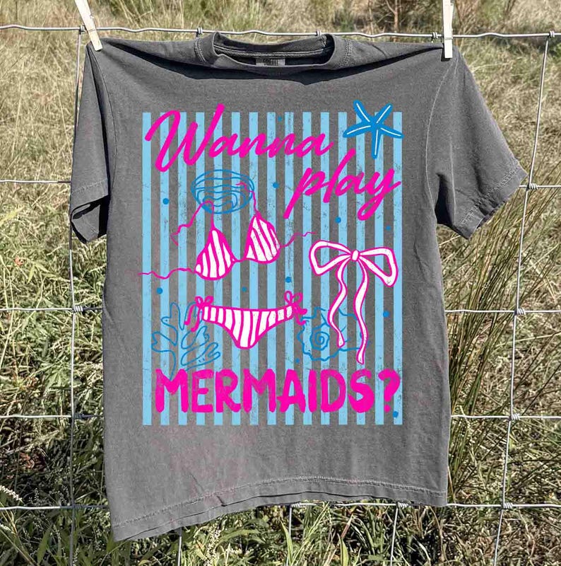 May include: Gray t-shirt with a graphic design featuring a bikini, bow, and starfish, with the text "Wanna Play Mermaids?" in pink and blue. The shirt is hanging on a clothesline.