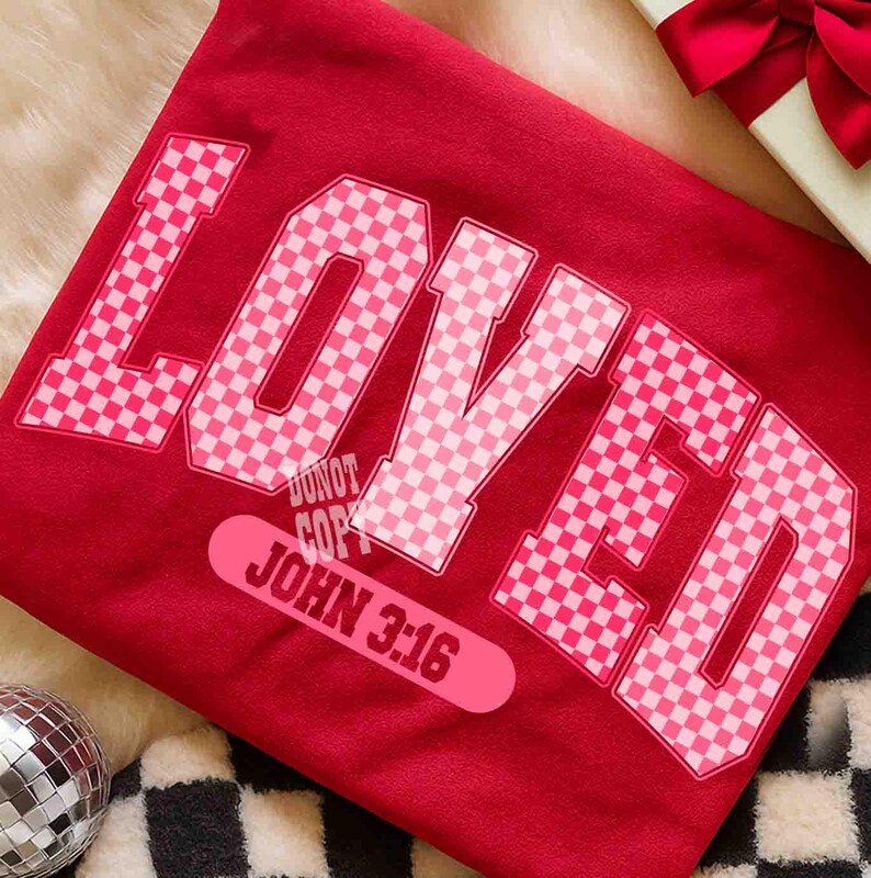 May include: A red t-shirt with the word "LOVED" in large, pink checkered letters. Below the word is a pink rectangle with the text "JOHN 3:16". A gift box with a red bow is in the background.