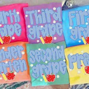以下が含まれることがあります： Fourth grade、Third grade、First grade、Kindergarten、Second grade、Pre-Kと書かれた青と白のギンガムチェック柄の文字が特徴の6枚のカラフルなTシャツ。それぞれに赤いリンゴと黄色い鉛筆のイラストが添えられています。