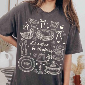 May include: Dark gray t-shirt with a white graphic design featuring teacups, plates, a duck, and the text "I'd rather be thrifting." The design includes floral and bow accents, creating a vintage aesthetic.