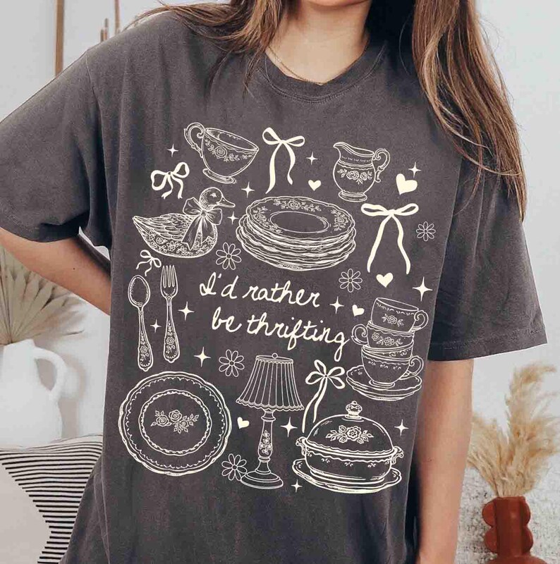 May include: Dark gray t-shirt with a white graphic design featuring teacups, plates, a duck, and the text "I'd rather be thrifting." The design includes floral and bow accents, creating a vintage aesthetic.