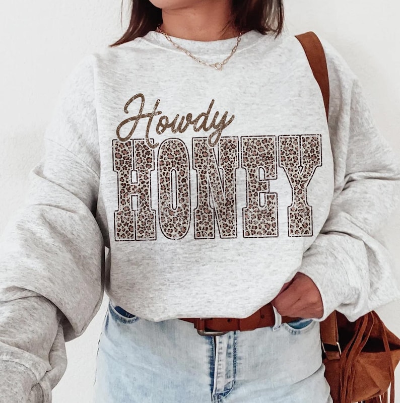 May include: A light gray sweatshirt with the words "Howdy HONEY" printed on the front. The word "Howdy" is in a script font, and "HONEY" is in a block letter font with a leopard print design. The sweatshirt has long sleeves.