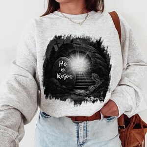 May include: Light gray sweatshirt featuring a black and white graphic of an open tomb with a bright light emanating from within. The text "He is Risen" is displayed on the front, along with the scripture reference Matthew 28.