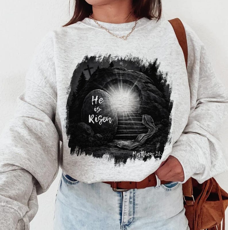 May include: Light gray sweatshirt featuring a black and white graphic of an open tomb with a bright light emanating from within. The text "He is Risen" is displayed on the front, along with the scripture reference Matthew 28.