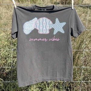 May include: Dark gray t-shirt with a seashell, scallop shell, and starfish graphic. The graphic is light blue, pink, and white. The text "summer vibes" is in pink script below the graphic. The shirt is hanging on a clothesline.