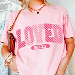 May include: Pink t-shirt with the word "LOVED" in a checkered pattern across the front. Below the word "LOVED" is the text "JOHN 3:16" in white on a pink rectangular background. The shirt is a solid pink color.