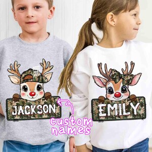 May include: Two children's sweatshirts, one gray and one white, each featuring a cartoon reindeer design. The reindeer wears a camouflage hat or bow and holds a sign with a custom name. The text "custom names" is also visible.