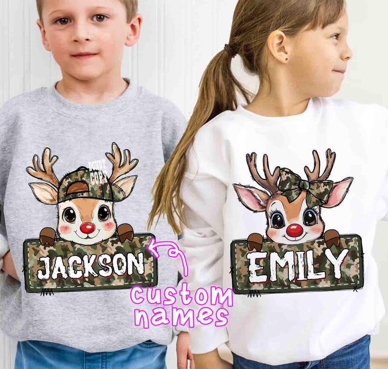 May include: Two children's sweatshirts, one gray and one white, each featuring a cartoon reindeer design. The reindeer wears a camouflage hat or bow and holds a sign with a custom name. The text "custom names" is also visible.