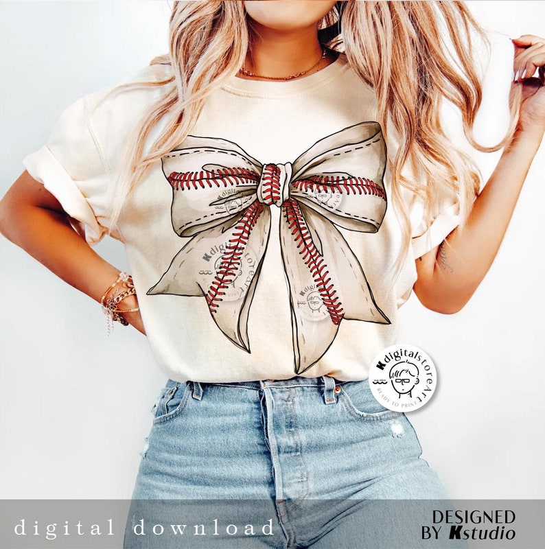 May include: A white t-shirt with a large brown bow made of baseballs with red stitching. The bow is centered on the shirt. The model is wearing blue jeans.
