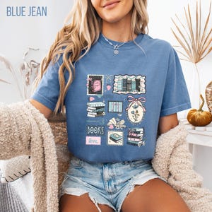 May include: Blue jean colored t-shirt with a graphic of postage stamps featuring books, flowers, and the word "BOOKS" in pink. The shirt has short sleeves and a relaxed fit.