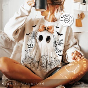 May include: Cream-colored sweatshirt featuring a ghost design with a peach bow, black bats, and spiderwebs. The ghost has large oval eyes and a lace-like bottom. The words "digital download" are at the bottom of the image.