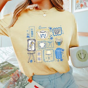 May include: Yellow t-shirt with a collage of vintage-style illustrations. The design includes a teapot, a plate, a lamp, and a chicken in a basket, all framed in blue. The word "Thrift" is also visible.