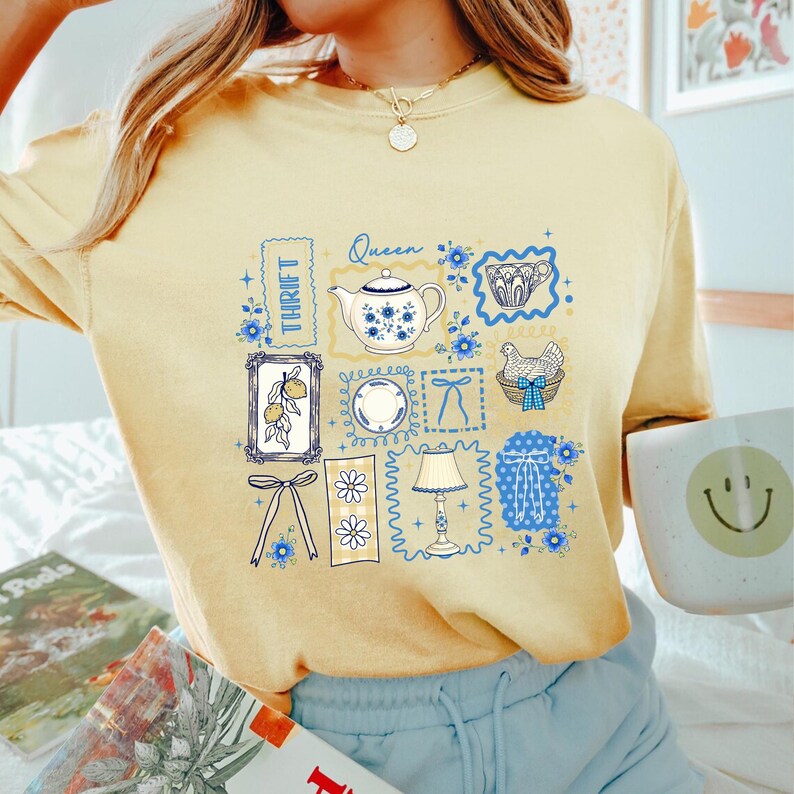 May include: Yellow t-shirt with a collage of vintage-style illustrations. The design includes a teapot, a plate, a lamp, and a chicken in a basket, all framed in blue. The word "Thrift" is also visible.