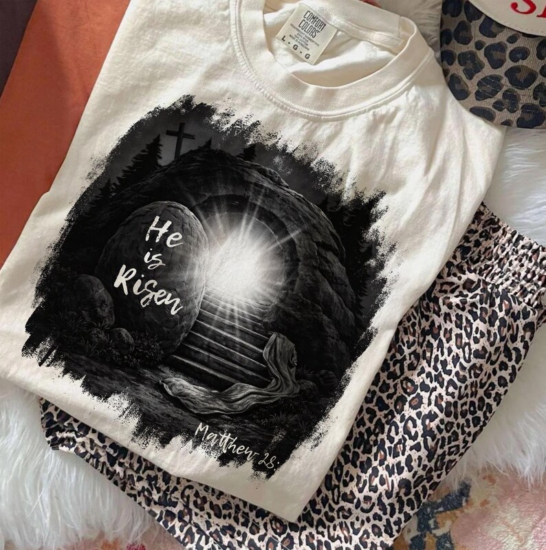 May include: Cream-colored t-shirt with a graphic of an open tomb and the text "He is Risen." The design is in black and white, with a bright light emanating from the tomb. The shirt is paired with leopard print pants.