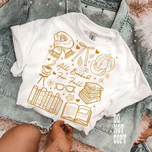 May include: White t-shirt with a gold graphic design featuring fall-themed illustrations, including a scarf, pumpkin, books, and the text "All Booked For Fall." The shirt is displayed on a denim jacket.