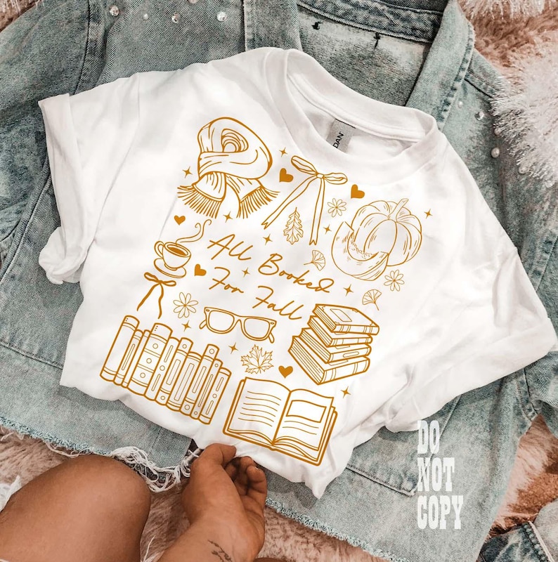 May include: White t-shirt with a gold graphic design featuring fall-themed illustrations, including a scarf, pumpkin, books, and the text "All Booked For Fall." The shirt is displayed on a denim jacket.