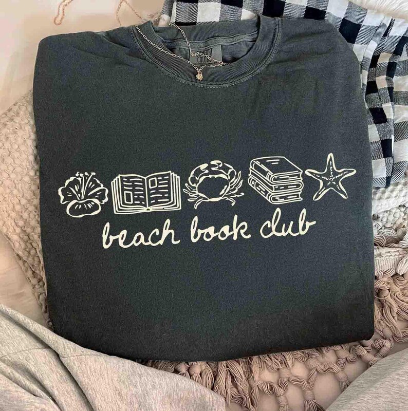 May include: Dark green sweatshirt with white graphic of a flower, open book, crab, stacked books, and starfish. The words "beach book club" are written below the graphics in a cursive font. A silver necklace with a small pendant is visible.