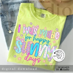 May include: A lime green t-shirt with the text "I was made for happy sunny days" in colorful, patterned letters. The shirt is folded on top of blue jeans, with a necklace and a hat in the background.