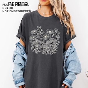 May include: Dark gray t-shirt featuring a white floral design. The shirt has a crew neck and short sleeves. The text "FLAT PEPPER, NOT 3D, NOT EMBROIDERED" is printed at the top left. The shirt is paired with a denim jacket.