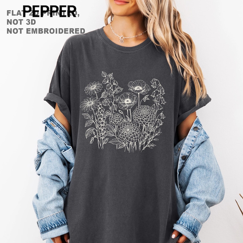 May include: Dark gray t-shirt featuring a white floral design. The shirt has a crew neck and short sleeves. The text "FLAT PEPPER, NOT 3D, NOT EMBROIDERED" is printed at the top left. The shirt is paired with a denim jacket.