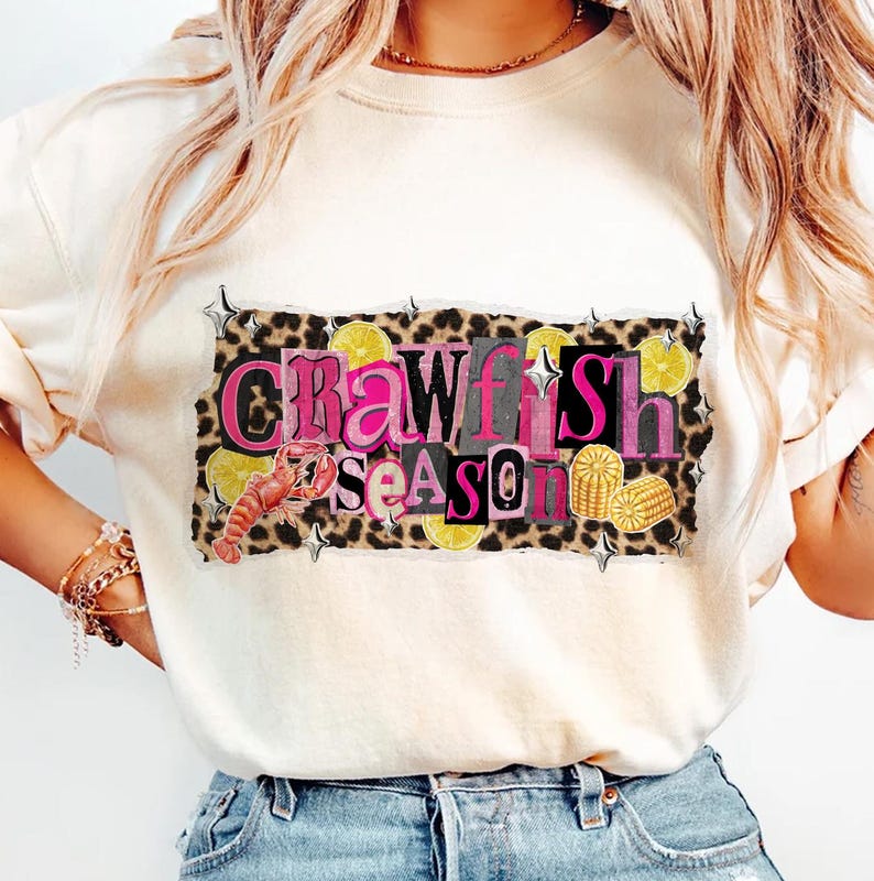 May include: Cream-colored t-shirt featuring a graphic that reads "Crawfish Season" in pink and black letters. The design includes a crawfish, lemons, and corn on a leopard print background. The shirt is a casual, everyday wear item.