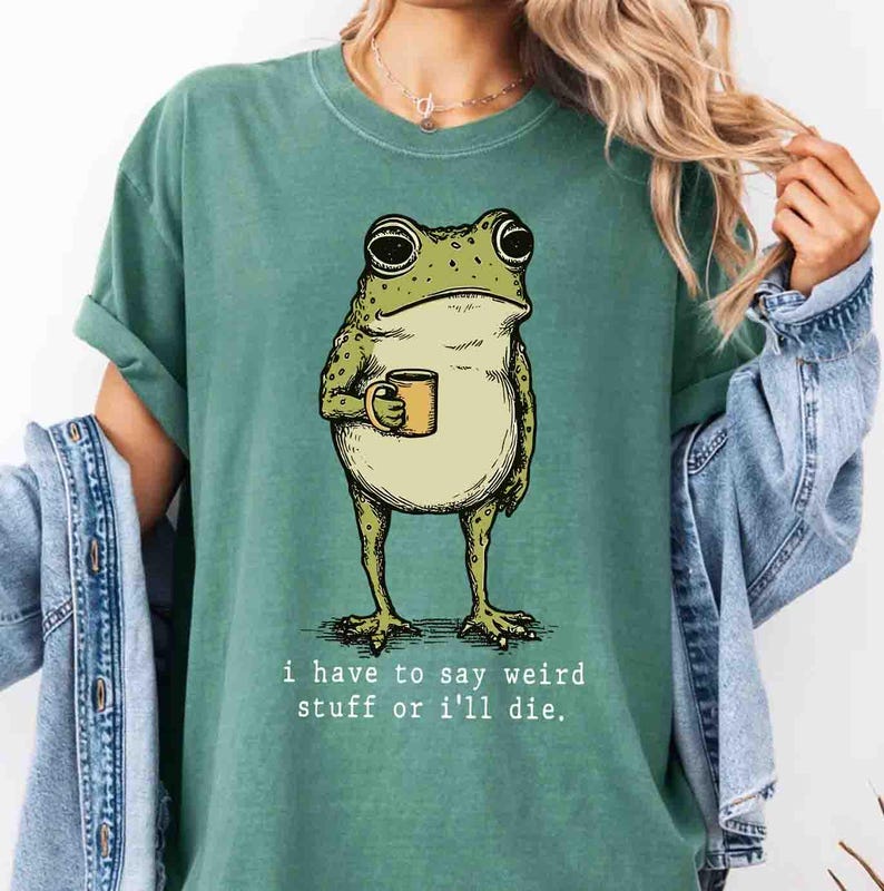 May include: Green t-shirt featuring a cartoon frog holding a yellow mug. The frog has a light green body with dark green spots. The text "I have to say weird stuff or I'll die" is printed below the frog. The shirt is a solid green color.