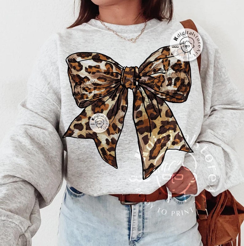 May include: A gray sweatshirt with a large brown and black leopard print bow on the front. The bow is tied in a knot and the ends are flowing down the front of the shirt.