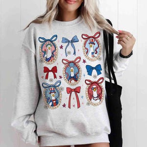 Op de afbeelding: Een lichtgrijs sweatshirt met een herhaald patroon van cartoonganzen in patriottische outfits, elk omlijst door een decoratieve strik. De ganzen dragen hoeden, vlinderdassen en zonnebrillen, met rode, witte en blauwe kleurenschema's.