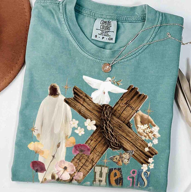 May include: A teal Comfort Colors t-shirt featuring a graphic of Jesus, a cross, and the words "He Is." The design includes a dove, flowers, and a butterfly. A silver necklace with a pendant is draped on the shirt.