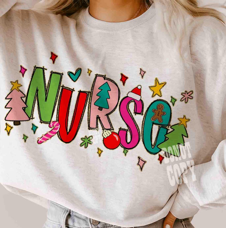 May include: A light gray sweatshirt with the word "NURSE" in colorful, festive lettering. The letters are decorated with Christmas-themed images like a Christmas tree, ornaments, and a gingerbread man. The sweatshirt has a crew neck.