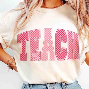 K&ouml;nnte beinhalten: Cremefarbenes Sweatshirt mit dem Wort "TEACH" in gro&szlig;en, rosa karierten Buchstaben auf der Vorderseite. Die Buchstaben haben einen Retro-College-Stil. Das Shirt ist ein l&auml;ssiges Kleidungsst&uuml;ck f&uuml;r den Alltag.