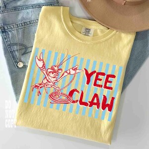 May include: Yellow t-shirt with a red and white cartoon crawfish wearing a cowboy hat, roping with the text "Yee Claw". The design is set against a blue striped background. The shirt is displayed with blue jeans and a tan hat.