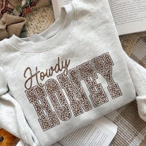 May include: A light gray sweatshirt with the words "Howdy HONEY" printed on the front. The word "Howdy" is in a brown script font, and "HONEY" is in a leopard print block font. The sweatshirt has long sleeves.