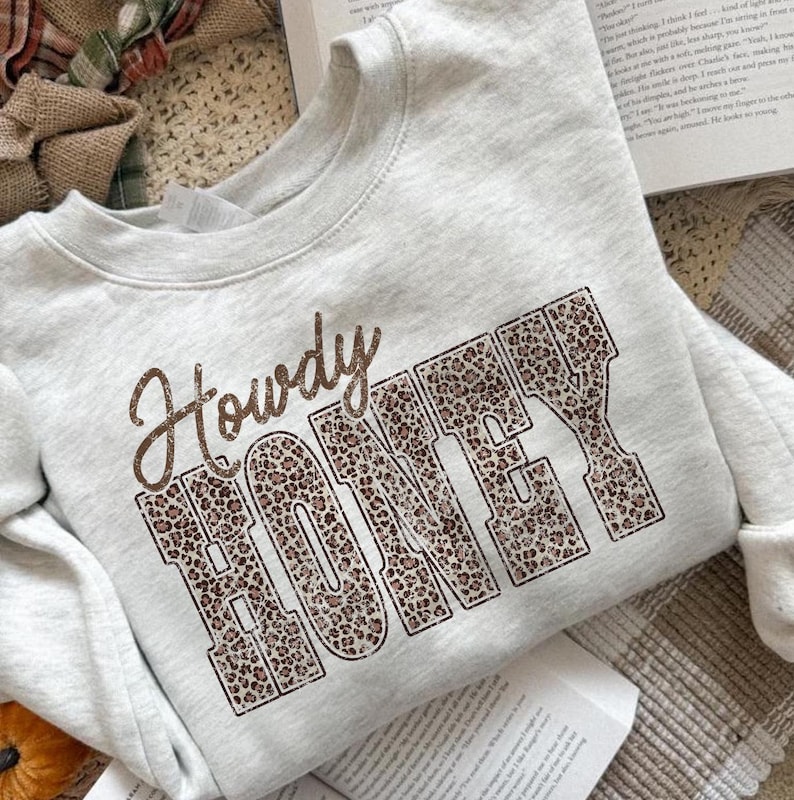May include: A light gray sweatshirt with the words "Howdy HONEY" printed on the front. The word "Howdy" is in a brown script font, and "HONEY" is in a leopard print block font. The sweatshirt has long sleeves.
