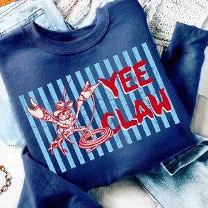 May include: Navy blue sweatshirt with a red and white graphic of a cowboy lobster roping with the text "YEE CLAW" in red. The design is set against a light blue striped background.